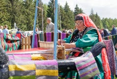 Starsza kobieta w Bashkir ubraniach sadza przy starym drewnianym oom i wyplata dywan Święto narodowe Sabantuy w miasto parku obraz stock Starsza kobieta w Bashkir ubraniach sadza przy starym drewnianym oom i wyplata dywan Święto narodowe Sabantuy w miasto parku obraz stock