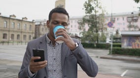 Steadicam tiró de hombre de negocios feliz joven usando smartphone y caminar con la taza de café al aire libre almacen de metraje de vídeo Steadicam tiró de hombre de negocios feliz joven usando smartphone y caminar con la taza de café al aire libre almacen de metraje de vídeo