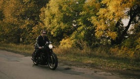 Stilfull ung man i solglasögon- och för läderomslag ridningmotorcykel på en asfaltväg på en solig dag i skog in lager videofilmer Stilfull ung man i solglasögon- och för läderomslag ridningmotorcykel på en asfaltväg på en solig dag i skog in lager videofilmer