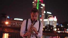 Street musician playing the saxophone on the waterfront on the background of night city. Beautiful night lights. stock video footage Street musician playing the saxophone on the waterfront on the background of night city. Beautiful night lights. stock video footage