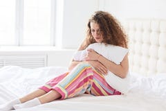 Stressed cury female wears pyjamas, holds pillow, suffers as has periods or menstruation, sits on soft bed with white bedclothes, royalty free stock image Stressed cury female wears pyjamas, holds pillow, suffers as has periods or menstruation, sits on soft bed with white bedclothes, royalty free stock image