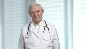 Stressed old angry physician having headache. Senior health care professional doctor with stethoscope. Blurred windows with louvers background Stressed old angry physician having headache. Senior health care professional doctor with stethoscope. Blurred windows with louvers background