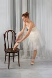 Studio shot of a calm beautiful ballerina in a white airy classic dress put her foot on a vienna chair and tying ribbons on pink royalty free stock photography Studio shot of a calm beautiful ballerina in a white airy classic dress put her foot on a vienna chair and tying ribbons on pink royalty free stock photography