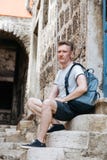 Stylish tourist. Man dressed in a white shirt and shorts with backpack over his shoulder. Sitting on the steps of European city. Stylish tourist. Man dressed in Royalty Free Stock Photos Stylish tourist. Man dressed in a white shirt and shorts with backpack over his shoulder. Sitting on the steps of European city. Stylish tourist. Man dressed in Royalty Free Stock Photos