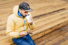 Stående av den stilfulla mannen med det bärande locket för skägg, det gula omslaget och jeans som dricker läckert kaffe och lyssn royaltyfri fotografi Stående av den stilfulla mannen med det bärande locket för skägg, det gula omslaget och jeans som dricker läckert kaffe och lyssn royaltyfri fotografi