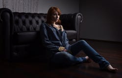 Stående av en rödhårig flicka i jeans skjorta och jeans som sitter på den svarta soffan på golvet royaltyfri foto Stående av en rödhårig flicka i jeans skjorta och jeans som sitter på den svarta soffan på golvet royaltyfri foto