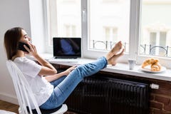 Stående av gladlynt sittingonstol för ung kvinna med ben på träfönsterbräda med frukosten som talar på telefonen arkivbild Stående av gladlynt sittingonstol för ung kvinna med ben på träfönsterbräda med frukosten som talar på telefonen arkivbild