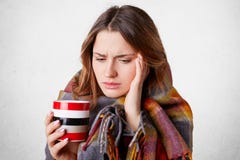 Ståenden av den härliga kvinnan har huvudvärk, har dålig förkylning, dricker varmt te, eller kaffe som slås in i rutig filt, ser arkivbild Ståenden av den härliga kvinnan har huvudvärk, har dålig förkylning, dricker varmt te, eller kaffe som slås in i rutig filt, ser arkivbild