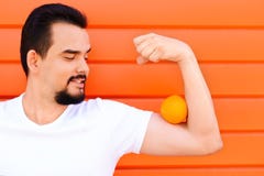 Ståenden av en le stilig man med mustaschen och skägget som håller en apelsin på hans biceps, tränga sig in mot den färgade vägge royaltyfri foto Ståenden av en le stilig man med mustaschen och skägget som håller en apelsin på hans biceps, tränga sig in mot den färgade vägge royaltyfri foto