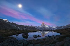 Summer landscape view of the Matterhorn reflecting in the water of the Stellisee lake. And the moon setting in a pink coloured sky at dawn