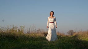 On a sunny day, a woman walks across a field in a white dress against a background of nature. stock video On a sunny day, a woman walks across a field in a white dress against a background of nature. stock video