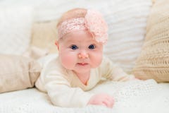 Sweet little baby with chubby cheeks and big blue eyes wearing white clothes and pink band with flower lying on bed royalty free stock photography Sweet little baby with chubby cheeks and big blue eyes wearing white clothes and pink band with flower lying on bed royalty free stock photography