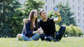Szczęśliwa potomstwo para opowiada w wideo gadce przez telefonu komórkowego, siedzi na trawie w parku i mówi z przyjaciółmi wideo zbiory Szczęśliwa potomstwo para opowiada w wideo gadce przez telefonu komórkowego, siedzi na trawie w parku i mówi z przyjaciółmi wideo zbiory