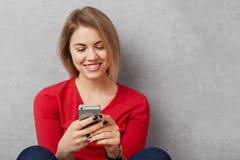 Szczęśliwa uśmiechnięta młoda kobieta w czerwonym pulowerze trzyma mądrze telefon i wiadomości online, zadowolony otrzymywać wiad zdjęcia royalty free Szczęśliwa uśmiechnięta młoda kobieta w czerwonym pulowerze trzyma mądrze telefon i wiadomości online, zadowolony otrzymywać wiad zdjęcia royalty free