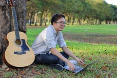 Szczęśliwy młody człowiek jest relaksujący z gitarą akustyczną w miasta lata parku i laptopem obraz stock Szczęśliwy młody człowiek jest relaksujący z gitarą akustyczną w miasta lata parku i laptopem obraz stock