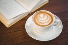 Tazza di cappuccino con arte del caffè accanto al libro aperto fotografia stock Tazza di cappuccino con arte del caffè accanto al libro aperto fotografia stock