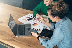 Teamwork. Two Young Business Women Sitting At Table In Front Of Laptop. On Table Is Tablet Computer And Paper Charts. Stock Photos Teamwork. Two Young Business Women Sitting At Table In Front Of Laptop. On Table Is Tablet Computer And Paper Charts. Stock Photos