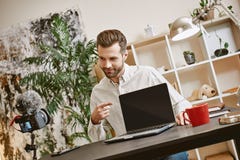 Technologia świat Pozytywnego młodego blogger magnetofonowy vlog i wskazywać na laptopie podczas gdy patrzejący kamerę fotografia stock Technologia świat Pozytywnego młodego blogger magnetofonowy vlog i wskazywać na laptopie podczas gdy patrzejący kamerę fotografia stock