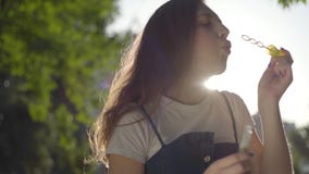 Teenage girl blowing soap bubbles at the camera in the park in the sun. Cute young woman spending time alone outdoors stock video footage Teenage girl blowing soap bubbles at the camera in the park in the sun. Cute young woman spending time alone outdoors stock video footage