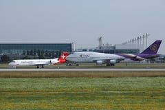 Thai Airways y Helvetic que llevan en taxi en el aeropuerto de Munich, MUC, vista delantera foto de archivo Thai Airways y Helvetic que llevan en taxi en el aeropuerto de Munich, MUC, vista delantera foto de archivo