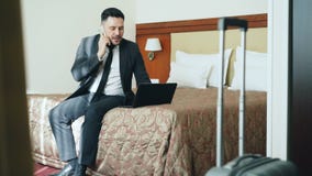 Tilt up of busy businessman in suit working on laptop and talking at mobile phone while sitting on bed in hotel room