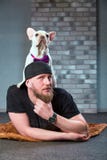 Tipo dei pantaloni a vita bassa con il suo bulldog francese piacevole dogdressed in cravatta a farfalla contro il muro di mattoni fotografia stock Tipo dei pantaloni a vita bassa con il suo bulldog francese piacevole dogdressed in cravatta a farfalla contro il muro di mattoni fotografia stock