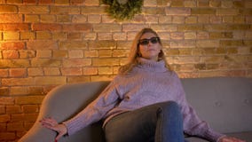 Tiro do close up da fêmea loura caucasiano adulta que olha um filme 3D na tevê nos vidros 3D ao sentar-se no sofá filme Tiro do close up da fêmea loura caucasiano adulta que olha um filme 3D na tevê nos vidros 3D ao sentar-se no sofá filme