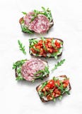 Tomatoes, Italian Sausage And Rocket Salad Whole Grain Bread Bruschetta On A Light Background, Top View. Copy Space Stock Photo Tomatoes, Italian Sausage And Rocket Salad Whole Grain Bread Bruschetta On A Light Background, Top View. Copy Space Stock Photo