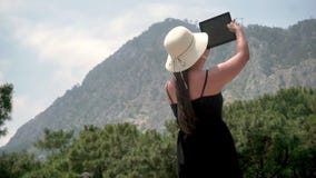 Tourist girl standing near a small green forest and a high hill, holding a gadget and taking pictures stock video footage Tourist girl standing near a small green forest and a high hill, holding a gadget and taking pictures stock video footage