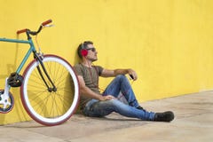 Tragende Sonnenbrille des jungen Mannes, die auf dem Boden und der hörenden Musik sich entspannt Sitzen gegen eine gelbe Wand mit lizenzfreies stockbild Tragende Sonnenbrille des jungen Mannes, die auf dem Boden und der hörenden Musik sich entspannt Sitzen gegen eine gelbe Wand mit lizenzfreies stockbild