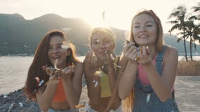 Trois femmes d'amis soufflant un pulvérisateur des confettis sur la plage au coucher du soleil banque de vidéos Trois femmes d'amis soufflant un pulvérisateur des confettis sur la plage au coucher du soleil banque de vidéos