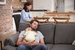 Trött fader With Baby Son sovande på Sofa As Mother Works Fotografering för Bildbyråer Trött fader With Baby Son sovande på Sofa As Mother Works Fotografering för Bildbyråer