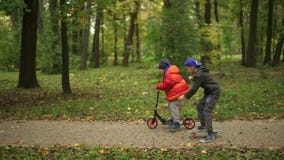 Twee tieners berijden een autoped en een stuiverraad Zij bewegen zich langs de weg in het park De herfstdag in het groene park, stock videobeelden Twee tieners berijden een autoped en een stuiverraad Zij bewegen zich langs de weg in het park De herfstdag in het groene park, stock videobeelden