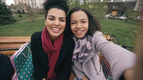 Two cheerful mixed race woman with shopping bags talking at video call with smartpone. Young girls sitting on bench have