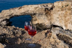 Two glasses of rose dry white wine served on rocks in blue sea bay with Love Bridge on background near Ayia Napa touristic town on