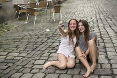 Two hilarious teenage girls are doing selfie on the phone sitting on the pavement. stock photo Two hilarious teenage girls are doing selfie on the phone sitting on the pavement. stock photo