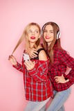 Two hipster woman posing, listening music by wireless headphones and making selfie by smartphone on the pink stock image Two hipster woman posing, listening music by wireless headphones and making selfie by smartphone on the pink stock image