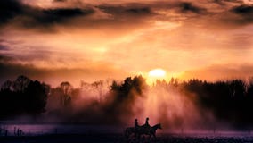 Two equestrians, horsemen horseriding at sunset on field. Sunbeams through trees, fog, orange clouds, pale violet shadows on snow.