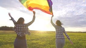 Two joyful young lesbian girls in shirts run across the field at sunset with an LGBT flag slow mo close up stock video footage Two joyful young lesbian girls in shirts run across the field at sunset with an LGBT flag slow mo close up stock video footage