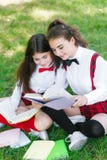 Two schoolgirls in school uniforms sit with books in the park. Schoolgirls or students are taught lessons in nature