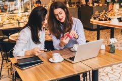 Two young happy women are sitting in cafe at table in front of laptop, using smartphone and laughing. Royalty Free Stock Photo Two young happy women are sitting in cafe at table in front of laptop, using smartphone and laughing. Royalty Free Stock Photo