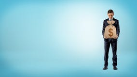 Um homem de negócios no fundo azul que guarda um saco do dinheiro de serapilheira com um sinal de dólar nele Fotografia de Stock Um homem de negócios no fundo azul que guarda um saco do dinheiro de serapilheira com um sinal de dólar nele Fotografia de Stock
