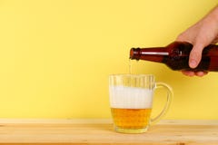 Um homem derrama a cerveja em uma caneca de uma garrafa em um fundo amarelo em uma tabela de madeira Copie o espaço foto de stock royalty free Um homem derrama a cerveja em uma caneca de uma garrafa em um fundo amarelo em uma tabela de madeira Copie o espaço foto de stock royalty free