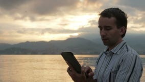 Um homem em uma camisa verifica mensagens na tabuleta durante o nascer do sol na praia do oceano Cores maravilhosas do céu vídeos de arquivo Um homem em uma camisa verifica mensagens na tabuleta durante o nascer do sol na praia do oceano Cores maravilhosas do céu vídeos de arquivo