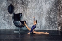 Uma ginasta da moça em um terno azul faz o esticão do exercício perto da cadeira contra a parede e os sorrisos cinzentos imagens de stock Uma ginasta da moça em um terno azul faz o esticão do exercício perto da cadeira contra a parede e os sorrisos cinzentos imagens de stock