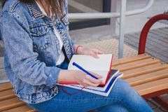 Uma menina na roupa das calças de brim senta-se em um banco, guarda-se cadernos e escreve-se Na rua, escreve em um caderno, estud foto de stock Uma menina na roupa das calças de brim senta-se em um banco, guarda-se cadernos e escreve-se Na rua, escreve em um caderno, estud foto de stock