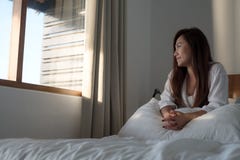 Uma mulher asiática bonita que senta-se na cama e que olha fora da janela fotografia de stock Uma mulher asiática bonita que senta-se na cama e que olha fora da janela fotografia de stock