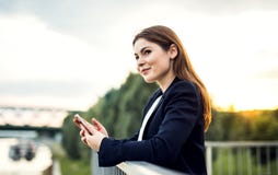 Uma mulher de negócios nova que está no banco de rio, usando o smartphone imagens de stock royalty free Uma mulher de negócios nova que está no banco de rio, usando o smartphone imagens de stock royalty free