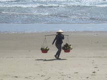 Uma mulher em um chapéu vietnamiano está andando ao longo de um Sandy Beach com as duas cestas do fruto imagens de stock Uma mulher em um chapéu vietnamiano está andando ao longo de um Sandy Beach com as duas cestas do fruto imagens de stock