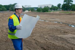 Un architecte dans un casque blanc, regardant un dessin d'un projet de construction, sur un terrain découvert, sol préparé photo libre de droits Un architecte dans un casque blanc, regardant un dessin d'un projet de construction, sur un terrain découvert, sol préparé photo libre de droits